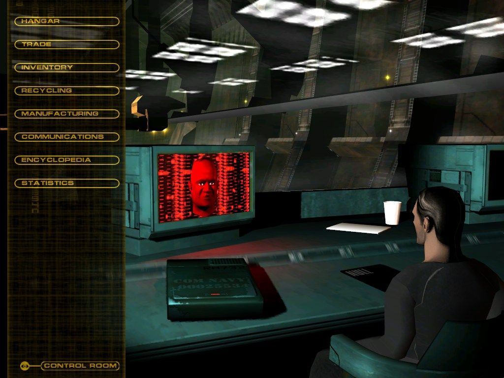 In a control room, someone is at a desk viewing Independence War 2: Edge Of Chaos on their monitor. The screen shows a face with a red background, along with menu options and tracking numbers on the left side.