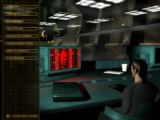 In a control room, someone is at a desk viewing Independence War 2: Edge Of Chaos on their monitor. The screen shows a face with a red background, along with menu options and tracking numbers on the left side.