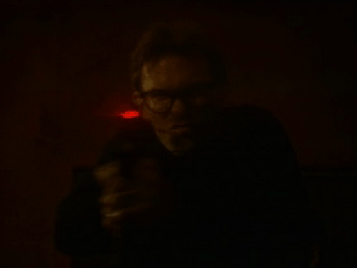 In a dimly lit environment with a red glow casting eerie shadows, a person wearing glasses aims a gun with the precision of delivering Johnny Mnemonic Disc 1 - 2 on Microsoft Windows PC CD.